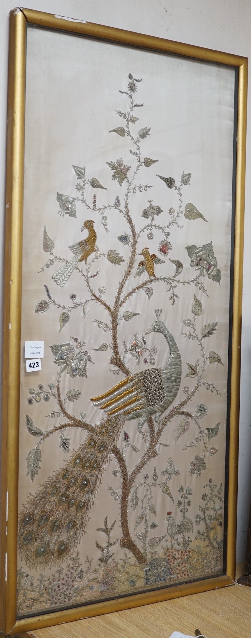 A framed silk, multi coloured metal thread embroidered Indian panel: a peacock seated amongst birds, flowers and leaves in a tree, 110cms x 51cms